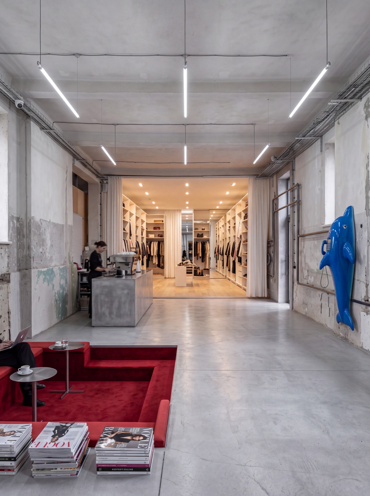 Inside a closet: a concrete hall under strip lighting with a coffee bar, a sunken red lounge stacked with magazines, and a lit room of rails and shelves at the far end.